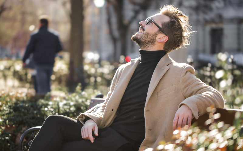 Hombre elegante con gafas de sol sentado en un banco mirando al cielo mientras sonríe
